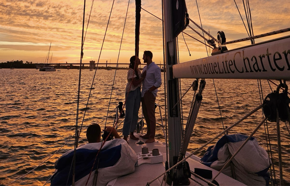 Couple enjoying a romantic moment on a sailboat during a vibrant Charleston SC sunset, with the Blue Life Charters logo visible and serene water in the background.